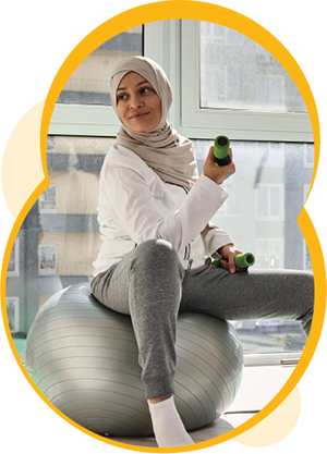 Une personne à la peau moyennement claire porte un hijab et est assise sur un ballon d’exercice. La personne tient des haltères dans ses mains. 