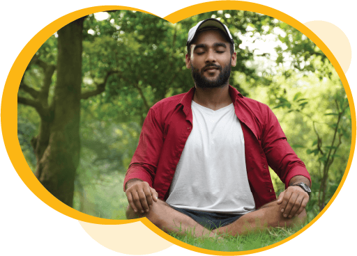 A person with medium-dark skin meditating while sitting on the ground outdoors, surrounded by grass and trees.