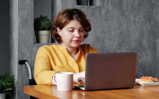 Une jeune femme dans un fauteuil roulant, travaillant sur un ordinateur portable, une tasse de café posée à coté