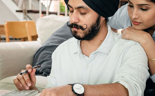 Un homme avec une moustache portant un turban tient un stylo et une calculatrice tandis qu'une femme regarde par-dessus son épaule.