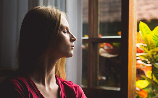 Une jeune femme, les yeux fermés, devant une fenêtre entre ouverte, qui médite
