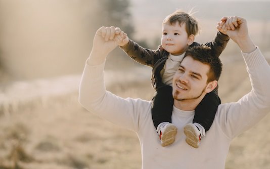 Un jeune papa avec son fils sur les épaules, le papa tenant les mains de son enfant dans les airs. 