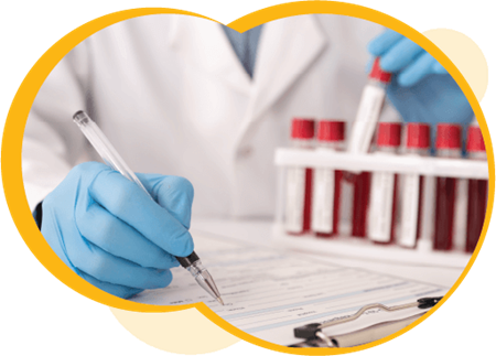 Un professionnel de la santé écrit sur une planchette à pince tout en analysant des échantillons de sang. Cette personne porte une blouse blanche et des gants d’examen bleus.