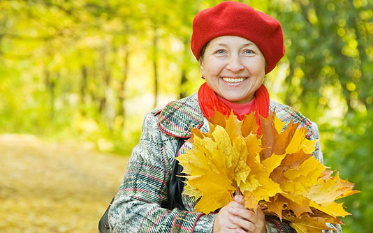 femme portant un beret rouge, tenant un bouquet de feuilles oranges dehors dans une forêt en automne. 