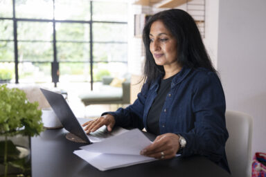 A person with medium skin sitting at a desk. They are using a laptop with one hand and holding a piece of paper in the other. Une personne &agrave; la peau moyennement fonc&eacute;e assise &agrave; un bureau. Elle utilise un ordinateur portable d&rsquo;une main et tient une feuille de papier dans l&rsquo;autre.